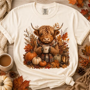 May include: A cream-colored short-sleeve t-shirt featuring a graphic of a fluffy brown Highland cow wearing a plaid scarf and holding a mug with a heart. The cow is surrounded by autumn leaves, pumpkins, and a lantern on a wooden surface.