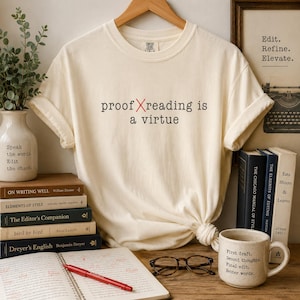 May include: A cream-colored short-sleeve t-shirt featuring the black text proof X reading is a virtue with a red X. The shirt is displayed with a stack of books, a red pen, a ceramic mug with text, and a plant in a white ceramic vase.