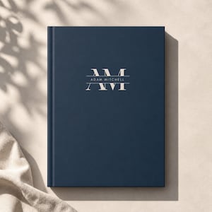 May include: A navy blue hardcover journal with the text "AM" above "Adam Mitchell" and "XVI" in a light color. The journal is closed and has a simple, elegant design. The background is a neutral color.