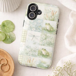 May include: A phone case with a patchwork design featuring frogs, dragonflies, and floral elements in shades of green and white. The case has a black camera cutout and a glossy finish, creating a nature-inspired aesthetic.