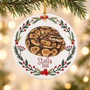 May include: A round white ceramic ornament featuring a coiled brown and tan ball python in the center. A wreath of green pine needles and red berries encircles the snake. The name Stella and the year 2026 are printed in red text below.