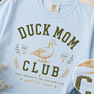 May include: Light blue t-shirt with a distressed green graphic that reads "DUCK MOM CLUB" with a duck illustration. The shirt also includes the text "EST. 2026" and "quack. love, repeat."
