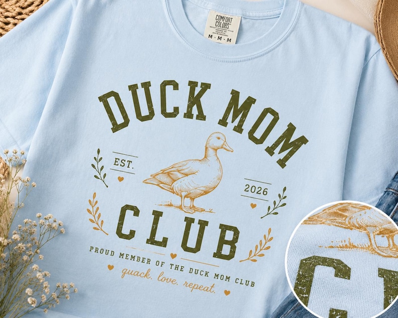 May include: Light blue t-shirt with a distressed green graphic that reads "DUCK MOM CLUB" with a duck illustration. The shirt also includes the text "EST. 2026" and "quack. love, repeat."
