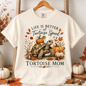 May include: An off-white short-sleeve t-shirt featuring a graphic of a tortoise with pumpkins on its back. The text reads "LIFE IS BETTER at Tortoise Speed" and "TORTOISE MOM" in black lettering, surrounded by autumn leaves and mushrooms.