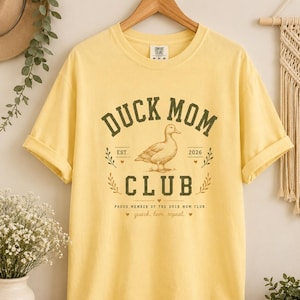May include: Yellow t-shirt with the text "DUCK MOM CLUB" in a green, vintage-style font. The design includes a duck illustration and the phrase "quack, love, repeat." The shirt is hanging on a wooden hanger.