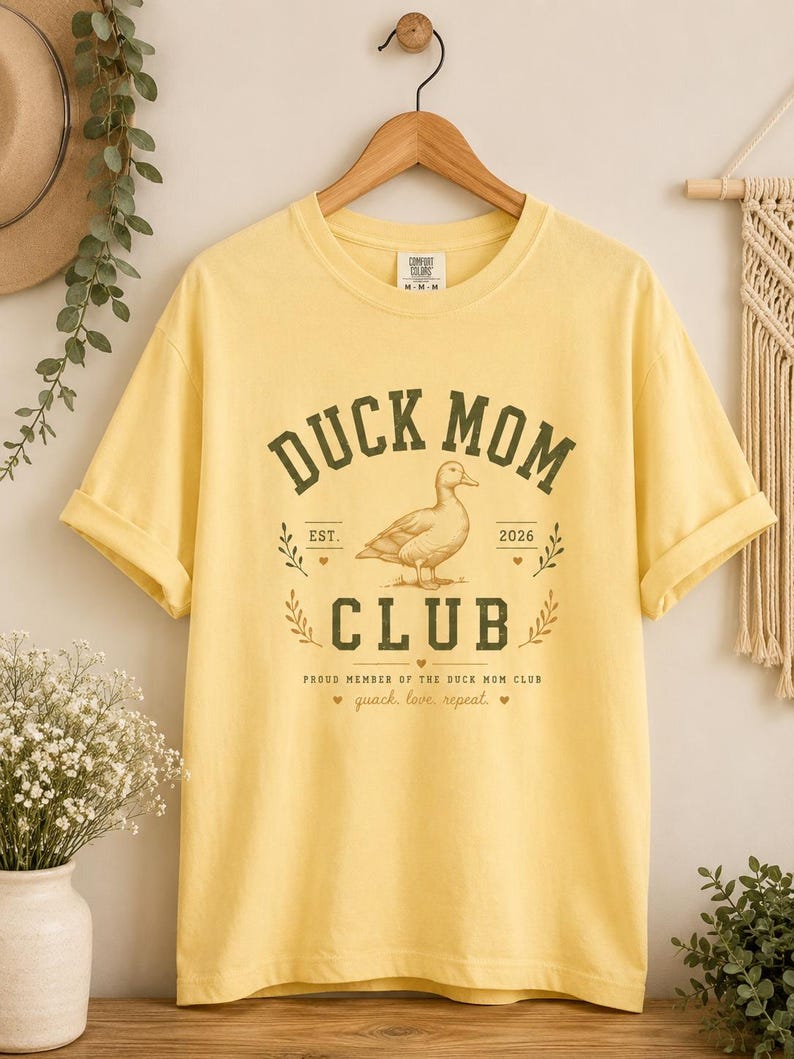 May include: Yellow t-shirt with the text "DUCK MOM CLUB" in a green, vintage-style font. The design includes a duck illustration and the phrase "quack, love, repeat." The shirt is hanging on a wooden hanger.