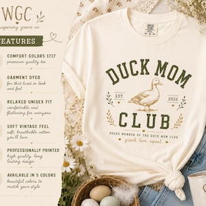 May include: Cream-colored t-shirt with a "Duck Mom Club" graphic in green and brown. The design includes a duck illustration and the words "quack, love, repeat." The shirt has a relaxed, unisex fit.