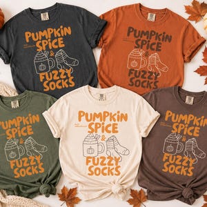May include: Five short-sleeve crew neck t-shirts in charcoal, burnt orange, olive green, cream, and dark brown. Each shirt features the text PUMPKIN SPICE & FUZZY SOCKS with a line art graphic of a mug and socks, printed in orange and white.