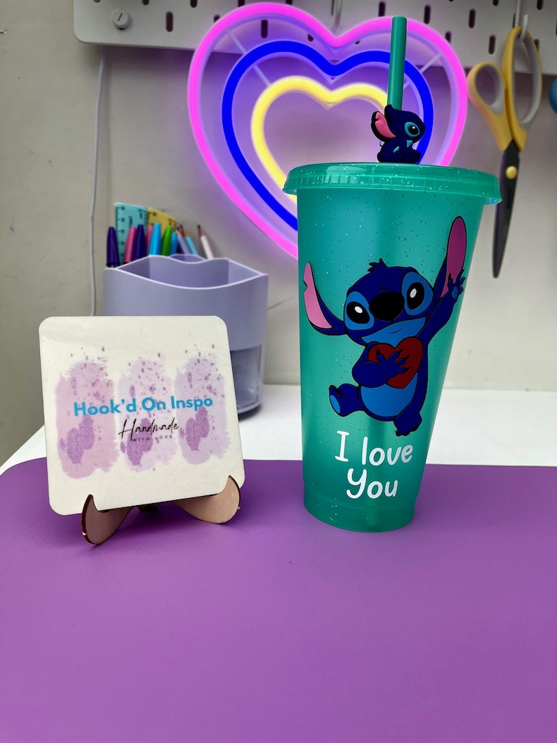 Puede incluir: Un vaso reutilizable azul turquesa con acabado brillante presenta un personaje azul Stitch sosteniendo un coraz&oacute;n rojo con el texto "I love you". El vaso tiene una pajita transparente con un personaje azul Stitch en la parte superior.