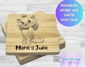 May include: A stack of square wooden coasters. The top coaster features an engraved image of a cartoon cat with a bow, and the words "Marie's Juice". The text "Personalised Natural Wood Coaster 10cm x 10cm" is in a circle in the background.