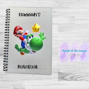 May include: A spiral-bound notebook with a frosted cover featuring a colorful graphic of Mario and Yoshi from the Super Mario video game series. The text "Hannah's" and "notebook" are printed in black.