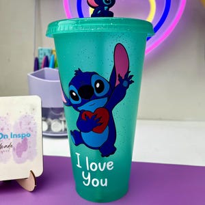 Puede incluir: Un vaso reutilizable azul turquesa con acabado brillante presenta un personaje azul Stitch sosteniendo un coraz&oacute;n rojo. El vaso tiene el texto "I love you" impreso en la parte delantera.