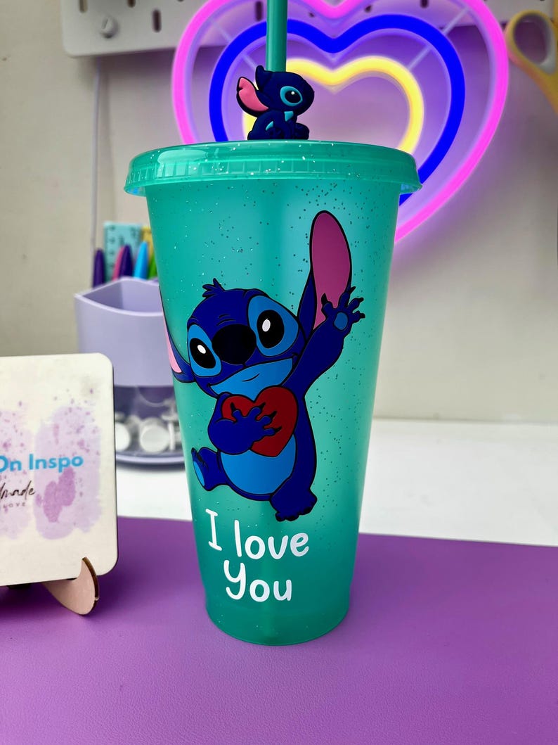 Puede incluir: Un vaso reutilizable azul turquesa con acabado brillante presenta un personaje azul Stitch sosteniendo un coraz&oacute;n rojo. El vaso tiene el texto "I love you" impreso en la parte delantera.