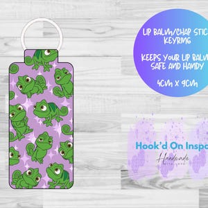 May include: A rectangular lip balm/chapstick keyring with a white metal ring. The keyring features a pattern of green cartoon chameleons on a light purple background. The text on the image says "Keeps your lip balm safe and handy". The keyring measures 4cm x 9cm.