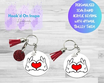 Puede incluir: Dos llaveros redondos blancos con un diseño de corazón rojo y manos enguantadas. Cada llavero tiene una borla roja y un pequeño dije redondo rojo brillante. El texto "Hook'd On Inspo Handmade" es visible.
