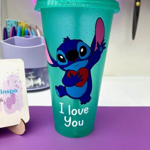 Puede incluir: Un vaso reutilizable azul turquesa con acabado brillante presenta un personaje de dibujos animados azul sosteniendo un coraz&oacute;n rojo. El personaje es Stitch de la pel&iacute;cula de Disney Lilo & Stitch. El texto "I love you" est&aacute; impreso debajo del personaje.