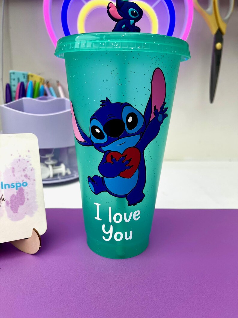 Puede incluir: Un vaso reutilizable azul turquesa con acabado brillante presenta un personaje de dibujos animados azul sosteniendo un coraz&oacute;n rojo. El personaje es Stitch de la pel&iacute;cula de Disney Lilo & Stitch. El texto "I love you" est&aacute; impreso debajo del personaje.