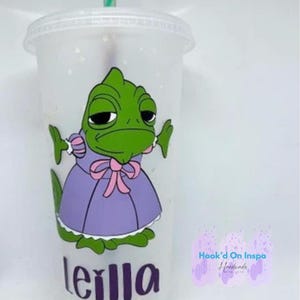 May include: A clear plastic tumbler with a green chameleon wearing a purple dress and a pink bow. The tumbler has the name "Leilla" written on it in purple letters. The tumbler is decorated with a purple and white splatter pattern. The text "Hook'd On Inspo Handmade" is visible in the bottom right corner.