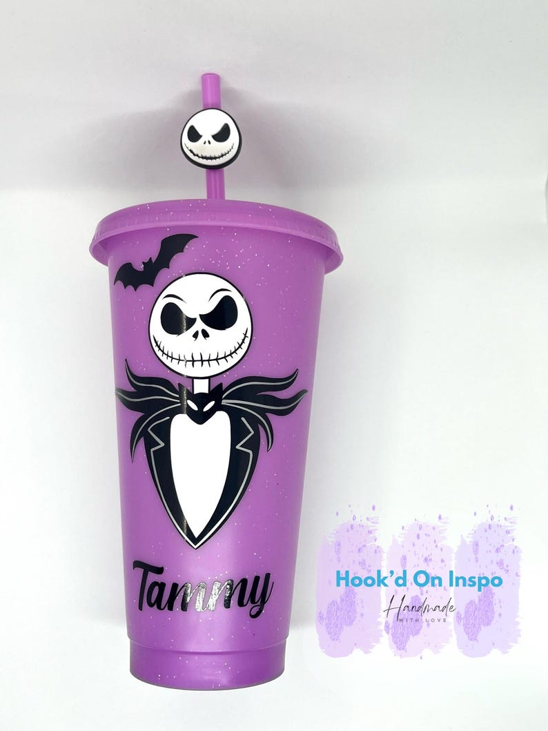 Puede incluir: Un vaso morado con un dise&ntilde;o de Jack Skellington, un murci&eacute;lago negro y el nombre "Tammy" en plata. El vaso tiene una pajita a juego y un adorno de Jack Skellington. El vaso est&aacute; decorado con purpurina.