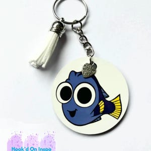 Könnte beinhalten: Ein weißer runder Schlüsselanhänger mit einem blauen Cartoon-Fisch mit großen Augen und einer gelben Schwanzflosse. Der Schlüsselanhänger hat einen silbernen Anhänger mit dem Text "Made with Love".