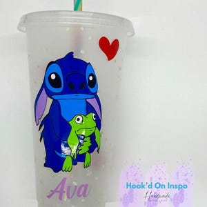 Puede incluir: Un vaso de pl&aacute;stico transparente con un personaje azul Stitch y una rana verde que sostiene un coraz&oacute;n rojo. El vaso tiene un acabado de purpurina morado y el nombre "Ava" est&aacute; escrito en la parte inferior. El vaso est&aacute; coronado con una pajita de rayas arco&iacute;ris. El texto "Hook'd On Inspo Handmade with Love" est&aacute; en el lateral del vaso.