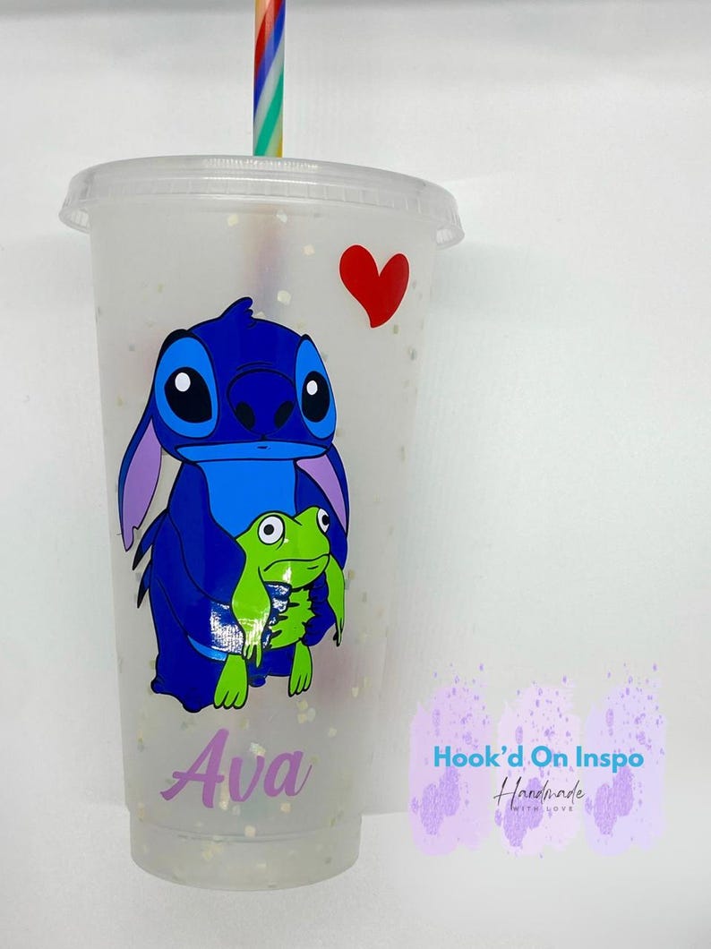 Puede incluir: Un vaso de pl&aacute;stico transparente con un personaje azul Stitch y una rana verde que sostiene un coraz&oacute;n rojo. El vaso tiene un acabado de purpurina morado y el nombre "Ava" est&aacute; escrito en la parte inferior. El vaso est&aacute; coronado con una pajita de rayas arco&iacute;ris. El texto "Hook'd On Inspo Handmade with Love" est&aacute; en el lateral del vaso.