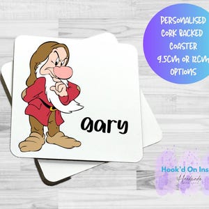 Op de afbeelding: Vierkante witte onderzetter met een cartoonafbeelding van Grumpy uit Sneeuwwitje, met de naam "Gary" in zwart. De onderzetter heeft een kurken achterkant en is verkrijgbaar in opties van 9,5 cm of 12 cm. Een paarse en blauwe cirkel en een paars logo zijn ook zichtbaar.