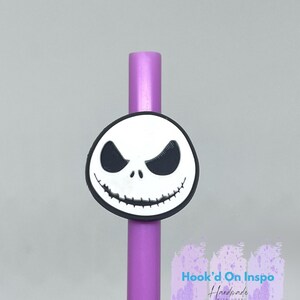 Puede incluir: Una pajita morada con un dije de la cara de Jack Skellington. El dije es blanco con ojos negros y un contorno negro. El texto "Hook'd On Inspo Handmade with love" est&aacute; visible.