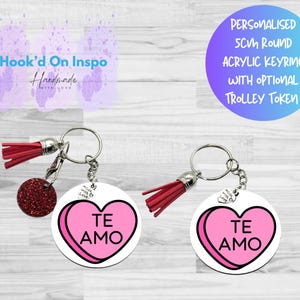 Op de afbeelding: Twee gepersonaliseerde ronde sleutelhangers met een roze en witte hartvormige snoepdesign met de tekst "TE AMO". Ze hebben rode kwastjes en een kleine, glinsterende rode bedel. De sleutelhangers zijn van acryl.