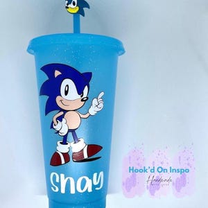Może przedstawiać: Jasnoniebieski kubek wielokrotnego użytku z błyszczącym wykończeniem. Kubek przedstawia grafikę Sonica the Hedgehog i napis "Shay" nadrukowany z przodu.