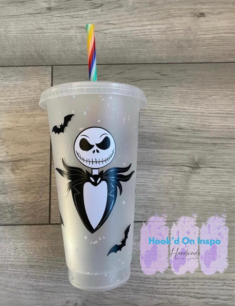 Puede incluir: Un vaso de pl&aacute;stico transparente con tapa y pajita a rayas arco&iacute;ris. El vaso presenta un gr&aacute;fico en blanco y negro de Jack Skellington de Pesadilla antes de Navidad, junto con siluetas de murci&eacute;lagos negros. El texto "Hook'd On Inspo Handmade With Love" es visible.