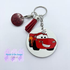 Può includere: Un portachiavi rosso di Saetta McQueen con un nappa rossa e un ciondolo rosso glitterato. Il portachiavi ha un portachiavi argentato e un piccolo ciondolo con la scritta "made with love".