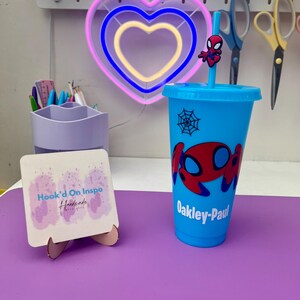 May include: A light blue plastic tumbler featuring a Spider-Man design with a matching straw and topper. The tumbler has the name "Oakley-Paul" printed on it. A neon heart-shaped light is in the background.