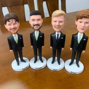 May include: Four bobblehead figures of men in black suits, each with a different hairstyle and facial features. Each figure stands on a white base with a name printed on it: David, Joseph, Dan, and Brian. The figures are on a wooden surface.