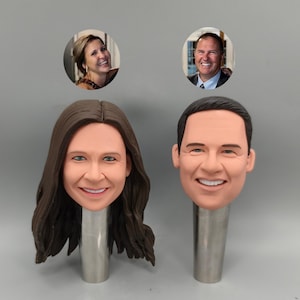 Puede incluir: Dos figuras personalizadas de bobblehead, una de una mujer con cabello largo castaño y la otra de un hombre con cabello corto negro. Cada figura tiene una cara sonriente y está montada sobre un soporte de metal plateado. Encima de cada bobblehead hay una pequeña foto de la persona.