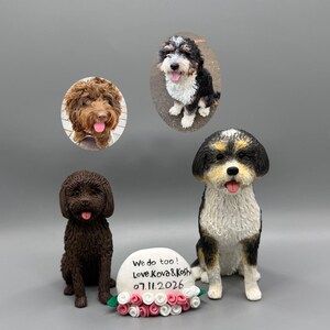 以下が含まれることがあります： 2体の犬のフィギュアが、「We do too! Love Kova & Koshi 07.11.2026」と書かれた白い石のプレートとピンクと白のバラの飾りの横に並んでいます。上部には、茶色の犬と黒・白・茶色の犬が写った2枚の楕円形の写真があります。