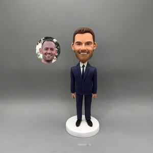 May include: A custom bobblehead of a man in a navy blue suit, white shirt, and black tie, standing on a white circular base. The head is a caricature with a smiling expression. A small photo of a man's face is in the background.