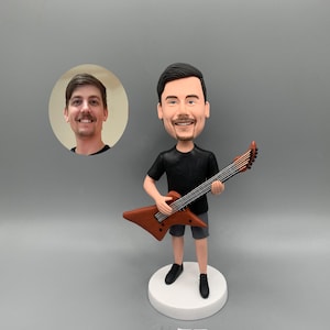 Pode incluir: Uma figura de bobblehead personalizada de um homem tocando uma guitarra elétrica marrom. A figura está vestindo uma camiseta preta, shorts cinza e sapatos pretos. Uma pequena foto do rosto do homem está no fundo. A figura está em uma base branca.