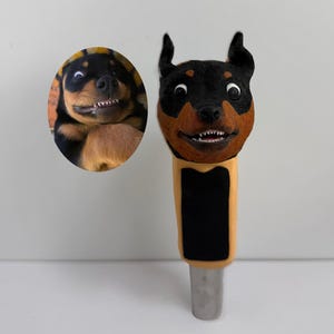 May include: A Rottweiler-themed tap handle with a smiling dog face, featuring black, brown, and tan colors. An oval image of a Rottweiler dog is next to the tap handle. The tap handle has a black rectangle and a silver metal base.