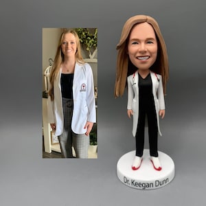 May include: A bobblehead figurine of a doctor in a white lab coat, black trousers, and red-trimmed shoes. The figurine has long brown hair and a stethoscope. The white base is inscribed with "Dr. Keegan Dunn". A photo of a woman in a white lab coat is in the background.