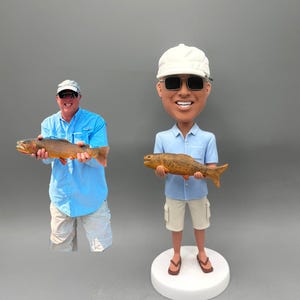 May include: A bobblehead figurine of a man wearing a white cap, sunglasses, and a light blue shirt, holding a brown fish. The figurine is standing on a white base. A photo of a man holding a fish is on the left.