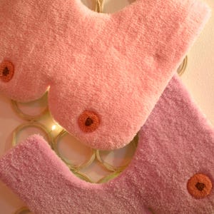 Può includere: Tre elementi decorativi in peluche rosa e lavanda, ciascuno con una forma arrotondata e un accento circolare arancione-marrone. Gli elementi sono disposti su uno sfondo di elementi circolari intrecciati e piccole luci.
