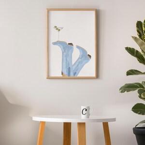 May include: A framed illustration of a pair of blue cowboy boots with a martini glass on top. The boots are upside down and the glass is filled with a green olive. The illustration is on a white background and is framed in a light wood frame. The frame is hanging on a white wall.