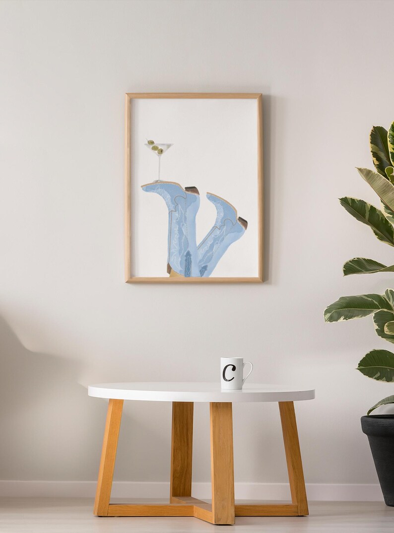 May include: A framed illustration of a pair of blue cowboy boots with a martini glass on top. The boots are upside down and the glass is filled with a green olive. The illustration is on a white background and is framed in a light wood frame. The frame is hanging on a white wall.