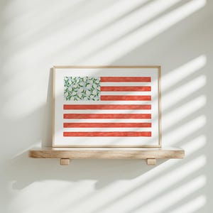 May include: A framed artwork depicting the American flag with a unique twist. The stripes are red, and the stars are replaced with a pattern of green leaves and white flowers. The artwork is displayed on a wooden shelf.