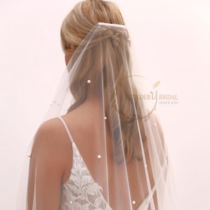 Op de afbeelding: Een witte bruids sluier met parelversiering. De sluier wordt gedragen door een persoon met blond haar, en de sluier is over de achterkant van het hoofd van de persoon gedrapeerd. De sluier is doorschijnend en heeft een delicate, romantische uitstraling.