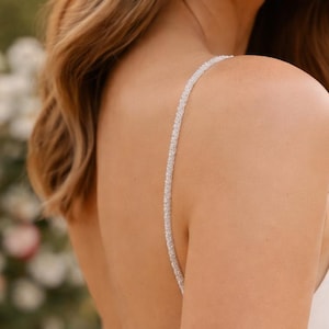 Puede incluir: Vestido de novia blanco con espalda baja y delicadas correas con cuentas. El vestido presenta un detalle de encaje en la cintura y una falda fluida. El cabello de la novia está peinado con ondas sueltas.