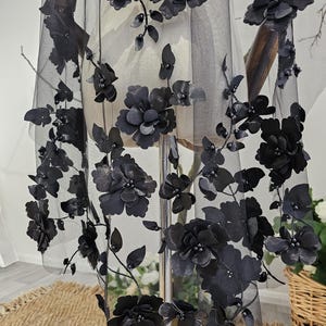 K&ouml;nnte beinhalten: Schwarzer floraler Schleier mit 3D-Blumen- und Bl&auml;tterverzierungen. Der Schleier besteht aus durchsichtigem schwarzem Stoff und weist mehrere gro&szlig;e, dunkle Bl&uuml;ten und kleinere Bl&auml;tter auf. Die Bl&uuml;ten sind geclustert und &uuml;ber den Schleier angeordnet.
