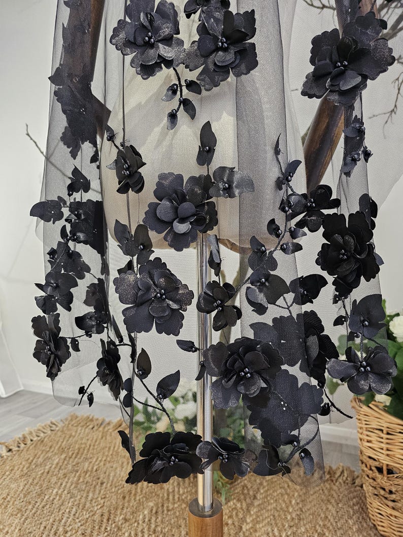 K&ouml;nnte beinhalten: Schwarzer floraler Schleier mit 3D-Blumen- und Bl&auml;tterverzierungen. Der Schleier besteht aus durchsichtigem schwarzem Stoff und weist mehrere gro&szlig;e, dunkle Bl&uuml;ten und kleinere Bl&auml;tter auf. Die Bl&uuml;ten sind geclustert und &uuml;ber den Schleier angeordnet.