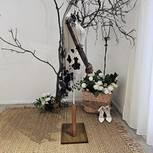 K&ouml;nnte beinhalten: Eine Schaufensterpuppe mit einem transparenten Schleier mit schwarzen Blumenstickereien. Die Schaufensterpuppe steht auf einem Holzsockel, mit einem Korb mit wei&szlig;en Blumen und einem Paar wei&szlig;er Abs&auml;tze. Die Szene spielt sich auf einem gewebten Teppich mit Fransen ab.